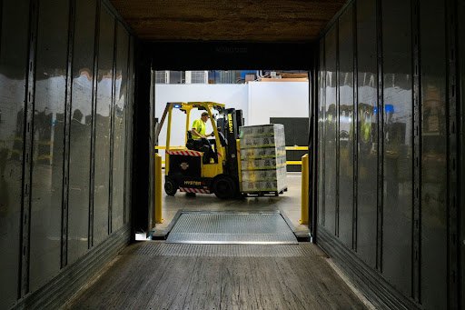 An operator drives a forklift carrying a pallet toward the rear entry of a semi truck