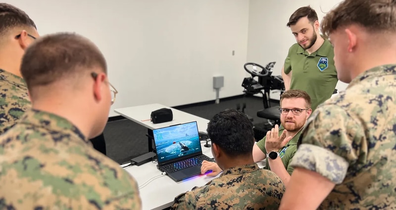 Two male instructors explain the XRT ACV training simulator to four male soldiers